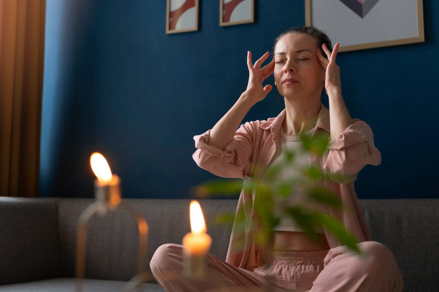 A woman seated calmly with eyes closed and fingertips at her temples in a softly lit room with candles.
