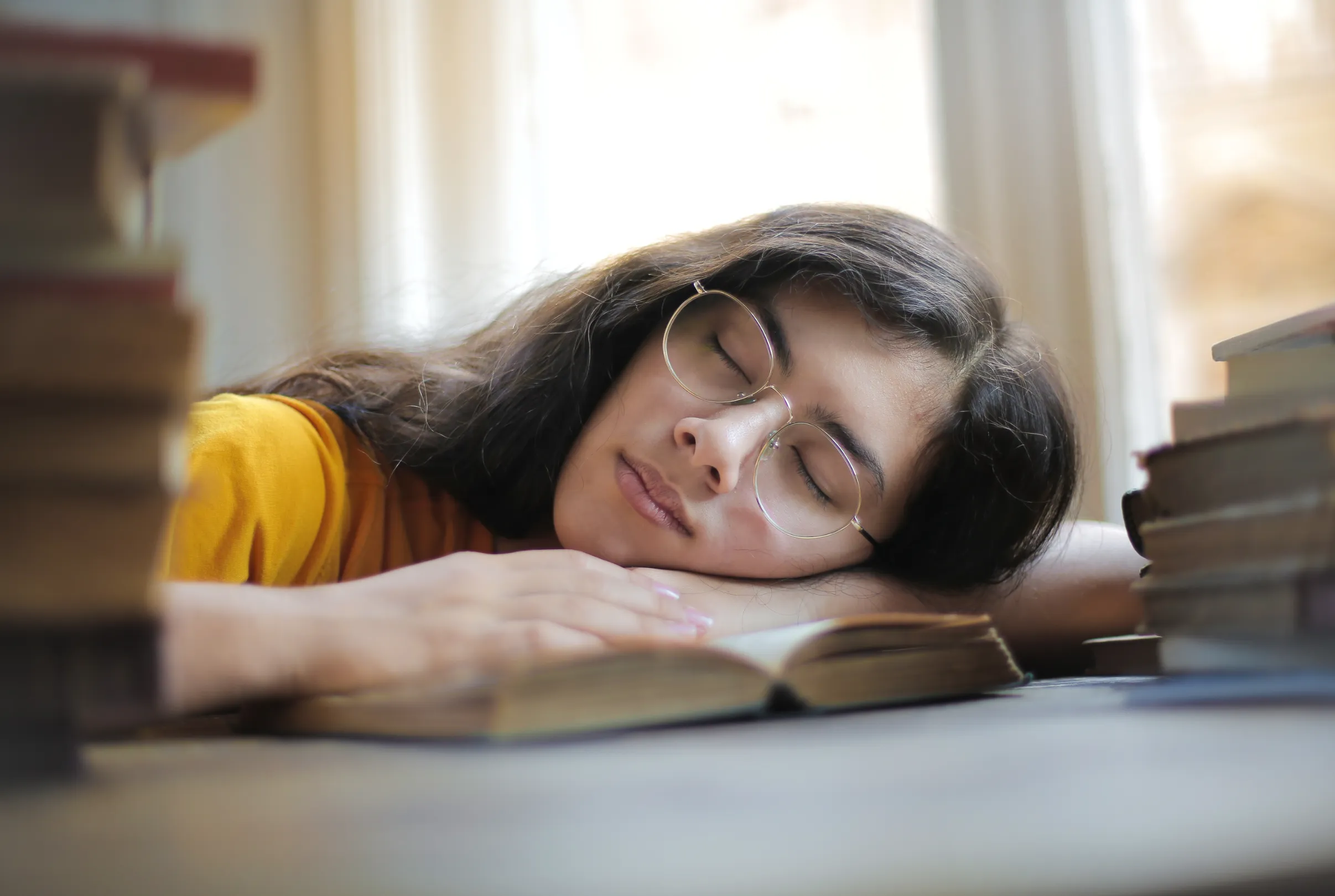 A young woman resting with her head on an open book at a desk, surrounded by stacks of books.