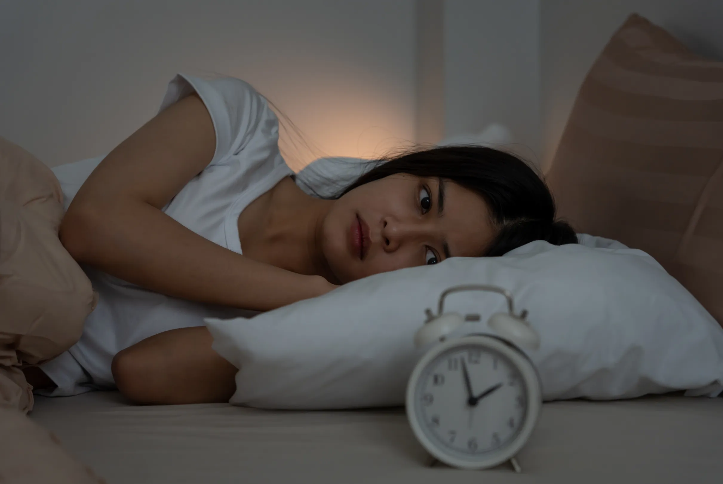 A woman awake in bed at night, eyes open, an alarm clock in the foreground.
