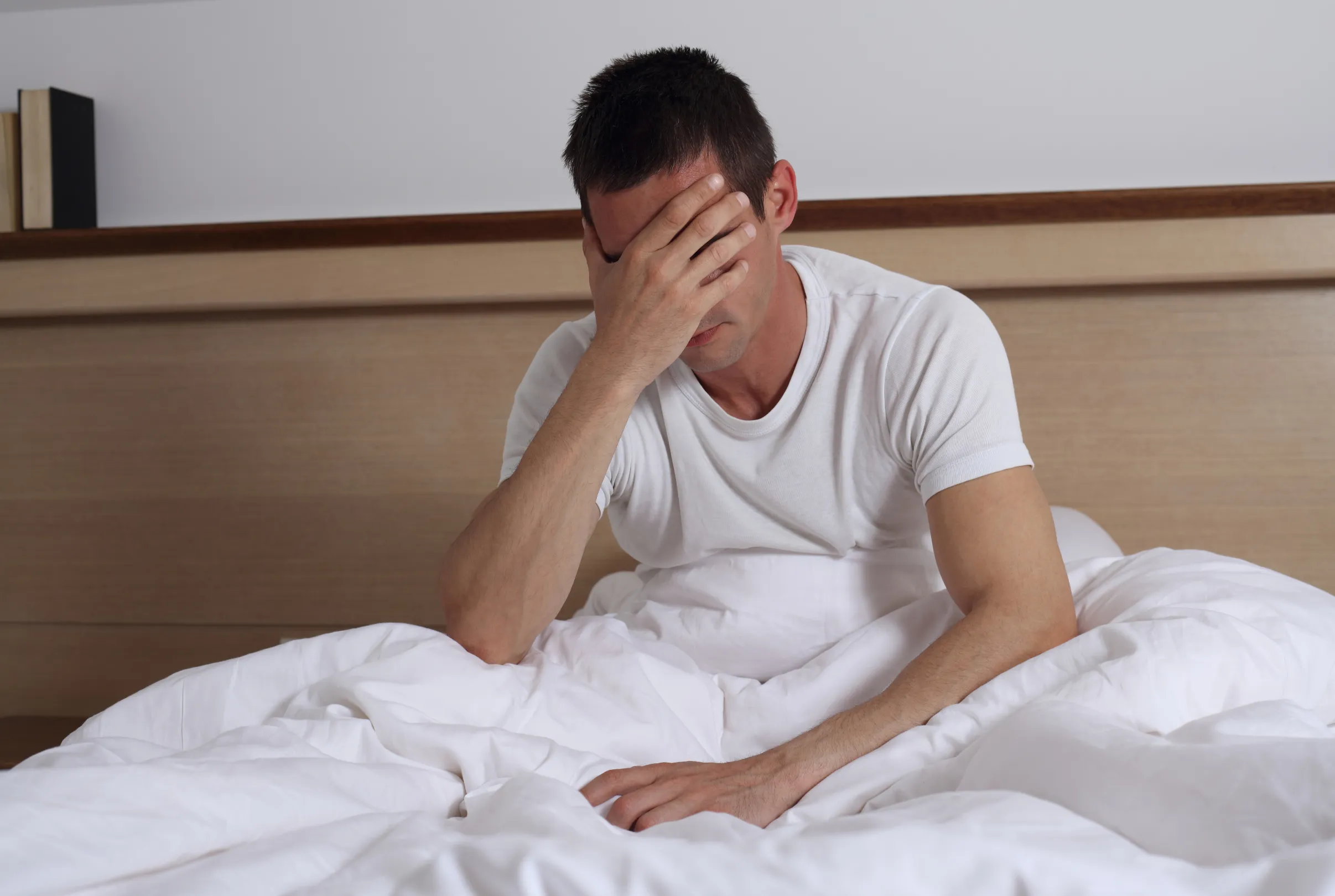 A man sitting up on the edge of his bed in the morning with one hand covering his face.